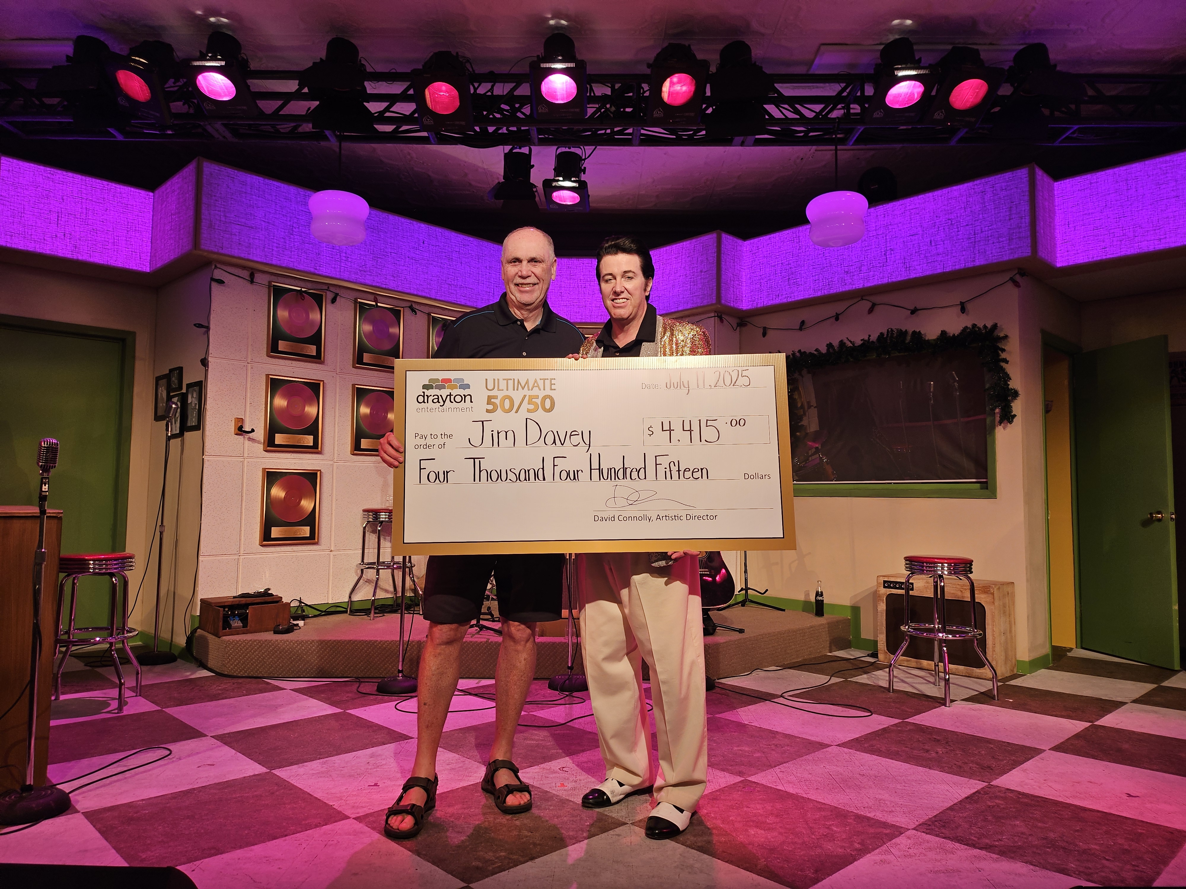 In this vibrant on-stage photo, Million Dollar Quartet actor Matt Cage, dressed as Elvis in a glittering gold jacket, presents a giant cheque to Jim Davey, the winner of Drayton Entertainment’s Ultimate 50/50 Draw. The cheque is made out for $4,415 and the two stand smiling in front of a retro set with pink and purple lighting and vintage gold records on the wall.